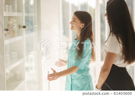 Female cosmetologis show to client cosmetology product. Doctor recommends different serum and face masks. Woman in cosmetoligy clinic choose feca balm Female cosmetologis show to client cosmetology product. Doctor recommends different serum and face masks. Woman in cosmetoligy clinic choose feca balm 61403600