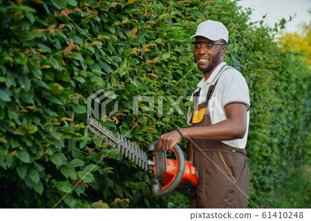 Cheerful afro gardener in uniform holding electronic trimmer Cheerful afro gardener in uniform holding electronic trimmer 61410248
