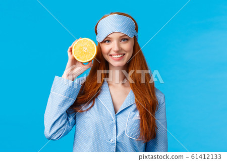 Start your morning right. Diet, healthy lifestyle and beauty concept. Cheerful glamour redhead woman in nightwear, holding slice orange near face, smiling, wearing sleep mask and pyjama 61412133