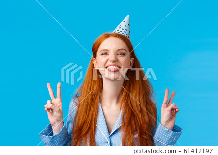Finally eighteen. Cheerful and enthusiastic carefree redhead woman with long hair, freckles wearing nightwear, b-day cap, showing tongue happy, make peace gestures, standing blue background 61412197