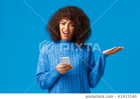 Wtf going on, someone explain me. Confused and frustrated bothered african-american woman in winter sweater, shrugging raise hand in dismay look questioned and pissed, holding smartphone 61413140