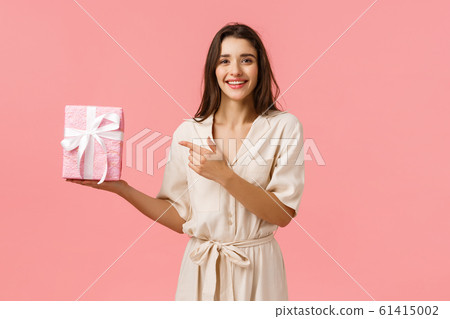 Girl prepared romantic gift for anniversary. Cheerful pretty young woman in cute dress holding gift box and pointing finger at present with delighted, happy smile, standing pink background 61415002