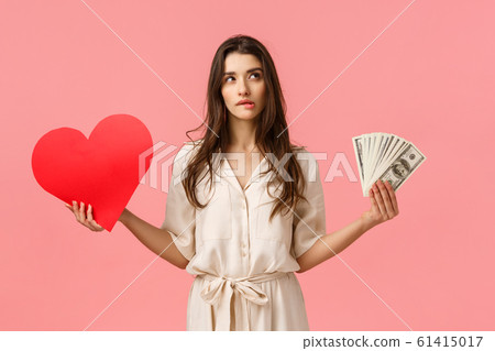 Romance, wealth and modern lifestyle concept. Alluring good-looking brunette female in dress, holding cash, money and love heart, biting lip look upper right corner unsure, making decision 61415017