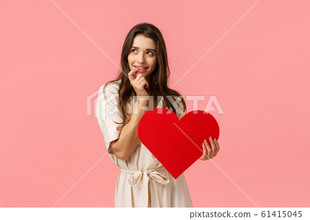 Coquettish and silly, dreamy cute, alluring brunette woman in dress, look left imaging perfect date, touching lip and smiling with tempting, eager expression, holding valentines heart, love concept 61415045