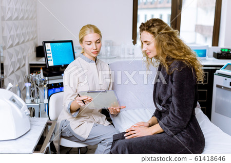 pretty cheerful blond woman in soft grey bathrobe sitting on daybed while female doctor cosmetologist talking about future beauty procedures. skin care, spa and cosmetology concept 61418646