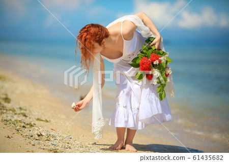 a red-haired girl collects seashells in a white dress on the beach with a bouquet of flowers a red-haired girl collects seashells in a white dress on the beach with a bouquet of flowers 61435682