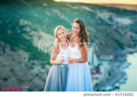 Two adult sisters women in dresses stand on the top of the mountain with glasses in their hands Two adult sisters women in dresses stand on the top of the mountain with glasses in their hands 61438882