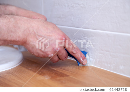 Closeup hands of professional plumber worker applying white sealant, joint compound, caulk to joint of wooden table top, beige tiled wall with rectangular tile using blue scraper 61442738