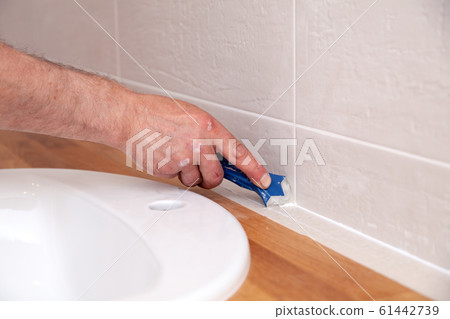 Closeup hands of professional plumber worker applying white sealant, joint compound, caulk to joint of wooden table top, beige tiled wall with rectangular tile using blue scraper 61442739