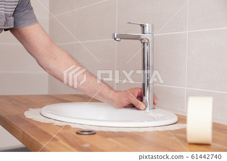 Closeup hands of a professional plumber worker installs a white oval ceramic sink on a wooden tabletop in the bathroom with beige tile, paste over sink with masking tape for applying sealant 61442740