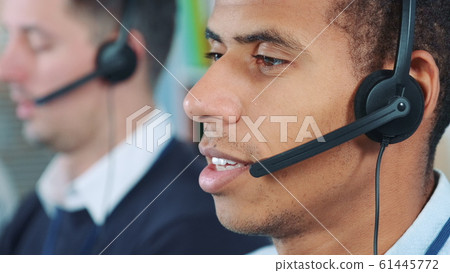 Close-up shot of mixed-race man in headset talking to somebody in office. In the background there is his colleague working. Close-up shot of mixed-race man in headset talking to somebody in office. In the background there is his colleague working. 61445772