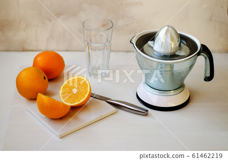 On a wooden table are two whole oranges and one cut in half. On the table lies a stone board and a knife. In the background is an empty glass cup. On the right side is a citrus juice squeezer. The On a wooden table are two whole oranges and one cut in half. On the table lies a stone board and a knife. In the background is an empty glass cup. On the right side is a citrus juice squeezer. The 61462219