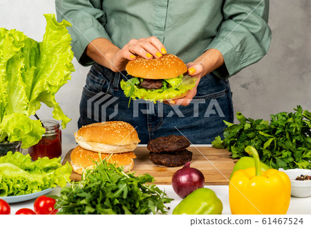 Woman is cooking tasty burger with cutlet, chesse and vegetables in the kitchen. Woman is cooking tasty burger with cutlet, chesse and vegetables in the kitchen. 61467524