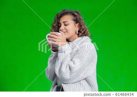 Sweet curly girl drinking hot cocoa standing against green background. Sweet curly girl drinking hot cocoa standing against green background. 61468434