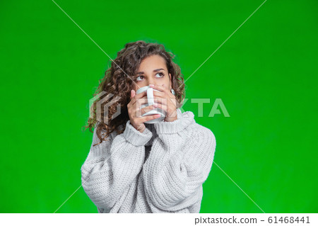 Young beautiful woman standing with cup of hot coffee wearing knitted warm sweater, dreaming about something inspiring. Young beautiful woman standing with cup of hot coffee wearing knitted warm sweater, dreaming about something inspiring. 61468441