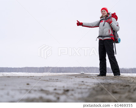 Woman hitch hiker try to catch a car showing thumb up on a road to share a ride. Winter time, front view, selected focus. Woman hitch hiker try to catch a car showing thumb up on a road to share a ride. Winter time, front view, selected focus. 61469355
