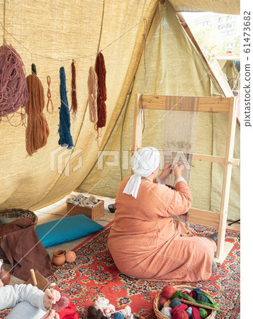 Times and Epochs Festival. Female Weaver Weaves a Canvas With a Pattern of Wool Yarn. Woman Weaves Using a Traditional Wooden Loom Times and Epochs Festival. Female Weaver Weaves a Canvas With a Pattern of Wool Yarn. Woman Weaves Using a Traditional Wooden Loom 61473682