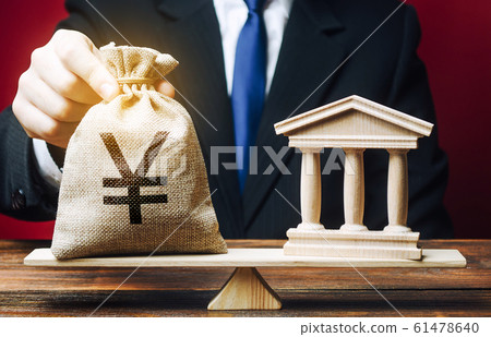 A man puts a Yen yuan money bag on scales opposite to building of government, bank, university. Budget and funding for normal functioning. Deposits and loans. Lobbying interests. Payment of taxes. 61478640