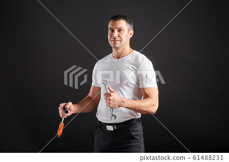 Athletic man with wrench on a black background. Not a typical worker, in a white t-shirt and trousers. Athletic man with wrench on a black background. Not a typical worker, in a white t-shirt and trousers. 61488231