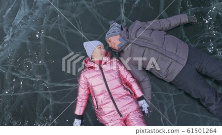 Couple has fun during winter walk against background of ice of frozen lake. Lovers lie on clear ice, have fun, kiss and hug. View from above. Couple has fun during winter walk against background of ice of frozen lake. Lovers lie on clear ice, have fun, kiss and hug. View from above. 61501264