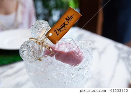 Beautiful crystal glass bottle with lid or stopper on ice crystal bowl with pink love or magical potion or liquor drink inside and vintage tag drink me from Alice in wonderland. Beautiful crystal glass bottle with lid or stopper on ice crystal bowl with pink love or magical potion or liquor drink inside and vintage tag drink me from Alice in wonderland. 61533520