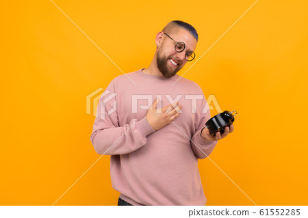 Young strong man with short black hair and glasses holds alarm clock and smiles Young strong man with short black hair and glasses holds alarm clock and smiles 61552285
