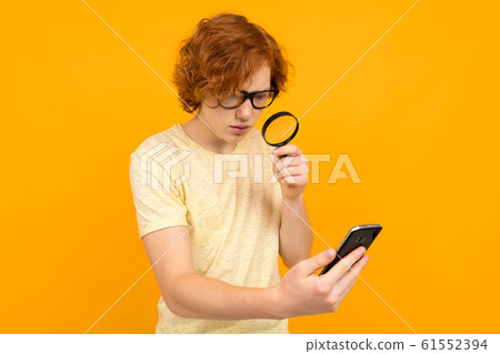 guy in glasses with a magnifier in his hand looks at a smartphone in his hand on a yellow background guy in glasses with a magnifier in his hand looks at a smartphone in his hand on a yellow background 61552394