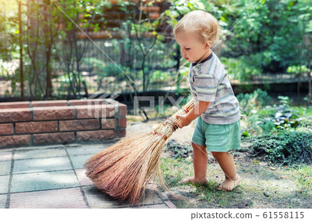 Cute adorable caucasian toddler boy playing holding broom at backyard in garden outdoors. Child little helper in t-short and shorts having fun sweeping and cleaning yard near house at countryside 61558115