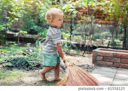 Cute adorable caucasian toddler boy playing holding broom at backyard in garden outdoors. Child little helper in t-short and shorts having fun sweeping and cleaning yard near house at countryside 61558125