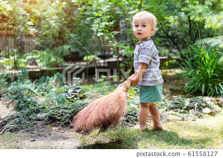 Cute adorable caucasian toddler boy playing holding broom at backyard in garden outdoors. Child little helper in t-short and shorts having fun sweeping and cleaning yard near house at countryside 61558127