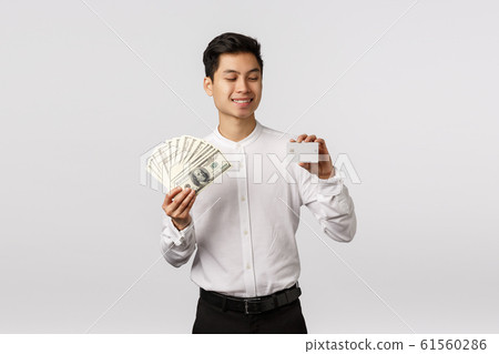 Finance, economy and business concept. Handsome asian businessman in formal outfit, holding cash and credit card, looking at banking payment method with pleased smile, spend money Finance, economy and business concept. Handsome asian businessman in formal outfit, holding cash and credit card, looking at banking payment method with pleased smile, spend money 61560286