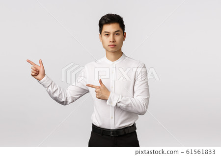 Serious-looking asian businessman give direction, pointing left, recommend visit site link, look corporate banner. Attractive elegant young man in white shirt and black pants advertising product 61561833