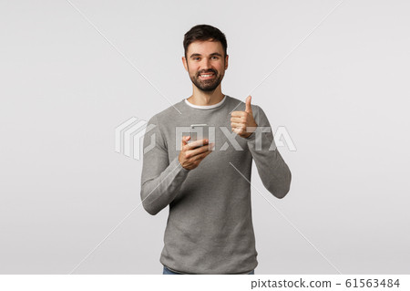 Nice, thanks for recommendation. Cheerful handsome bearded man in grey sweater holding smartphone, make thumb-up gesture, like app, advertise application, approve device or online shop Nice, thanks for recommendation. Cheerful handsome bearded man in grey sweater holding smartphone, make thumb-up gesture, like app, advertise application, approve device or online shop 61563484