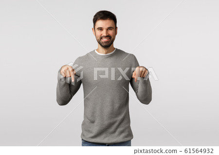 Time to see it, look here. Happy, charismatic friendly-looking smiling bearded male in grey sweater, invite event, share link, discuss promo offer, pointing down to promote product, white background 61564932