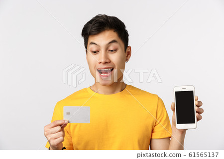 Close-up shot enthusiastic guy decided put money deposit, get credit to pay for trip, holding smartphone showing display and banking card, smiling amused, standing white background Close-up shot enthusiastic guy decided put money deposit, get credit to pay for trip, holding smartphone showing display and banking card, smiling amused, standing white background 61565137