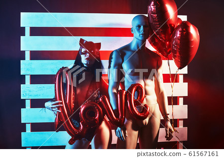 Sexy woman wearing a black cat mask standing hugging mannequin and holding heart-shaped balloons and a love sign and looks very sensually, Valentine's day concept 61567161