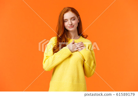 Tender and romantic, lovely young modern redhead woman 20s, touch heart and close eyes, remember dearest, touching moment, recall perfect date, keep memories in soul, orange background 61589179