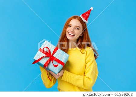 Holidays, celebration and presents concept. Cheerful redhead girl love christmas, enjoy celebrating with friends, receive gift in wrapped box from secret santa, wearing cute hat, blue background 61589268