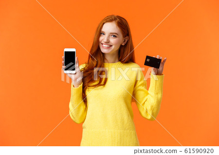 Redhead caucasian girl showing application on smartphone as holding mobile phone and credit card, using mobile banking app, cheerfully smiling camera, advertising over orange background 61590920