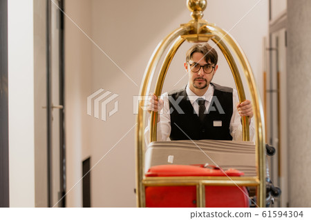 Young elegant porter in eyeglasses pushing cart with luggage of hotel guests Young elegant porter in eyeglasses pushing cart with luggage of hotel guests 61594304