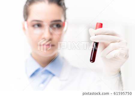 Hand of young female clinician in gloves holding flask with blood test Hand of young female clinician in gloves holding flask with blood test 61595750