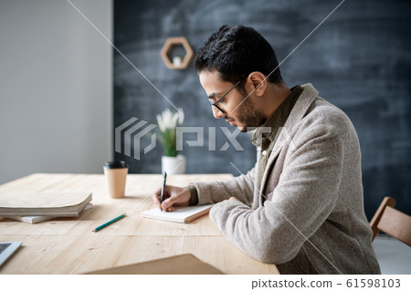 Young casual student making notes in notebook while sitting by wooden table Young casual student making notes in notebook while sitting by wooden table 61598103