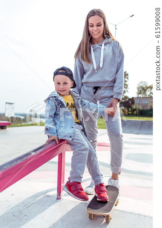 Young family little boy 3-5 years old, mother woman, having fun, smiling, rejoice, learning to ride a skateboard, in the summer in a city park. Weekend rest on sports field. Skateboard casual wear. Young family little boy 3-5 years old, mother woman, having fun, smiling, rejoice, learning to ride a skateboard, in the summer in a city park. Weekend rest on sports field. Skateboard casual wear. 61599008