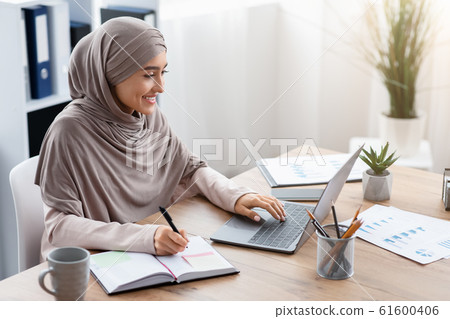 Muslim woman in hijab using laptop and taking notes in office Muslim woman in hijab using laptop and taking notes in office 61600406