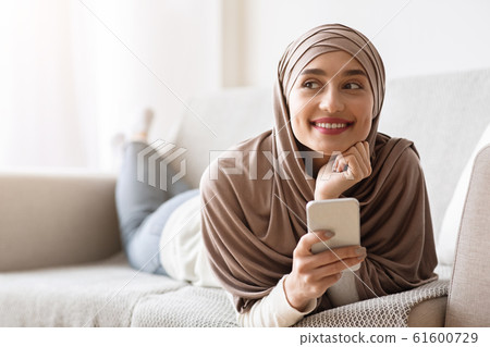Dreamy arabic girl in headscarf lying on couch with smartphone 61600729