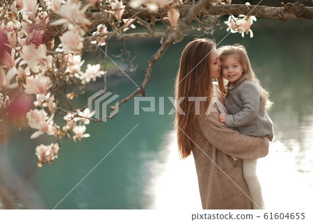 A woman holds a child in her arms against the background of a lake in a garden with magnolia A woman holds a child in her arms against the background of a lake in a garden with magnolia 61604655
