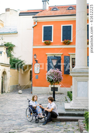 Pretty young smiling woman in the wheelchair and handsome man sitting on the stone stairs, looking each other in love, walking in the old city center. Bright orange building on the background 61612225