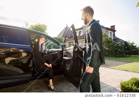 Businessman coming to car and opens door for young woman, talking phone. Businesswoman get out from the auto and is ready to go with her colleague to office building Businessman coming to car and opens door for young woman, talking phone. Businesswoman get out from the auto and is ready to go with her colleague to office building 61612336