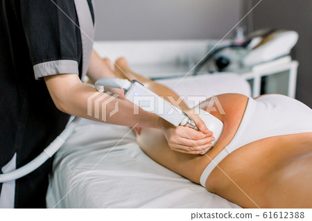 Female doctor cosmetologist doing roller massage with apparatus on female client thighs, close up. Roller massage in medical spa center 61612388