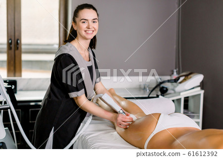 Young smiling woman cosmetologist with roller doing buttocks massage for her female client in modern spa medical center. 61612392